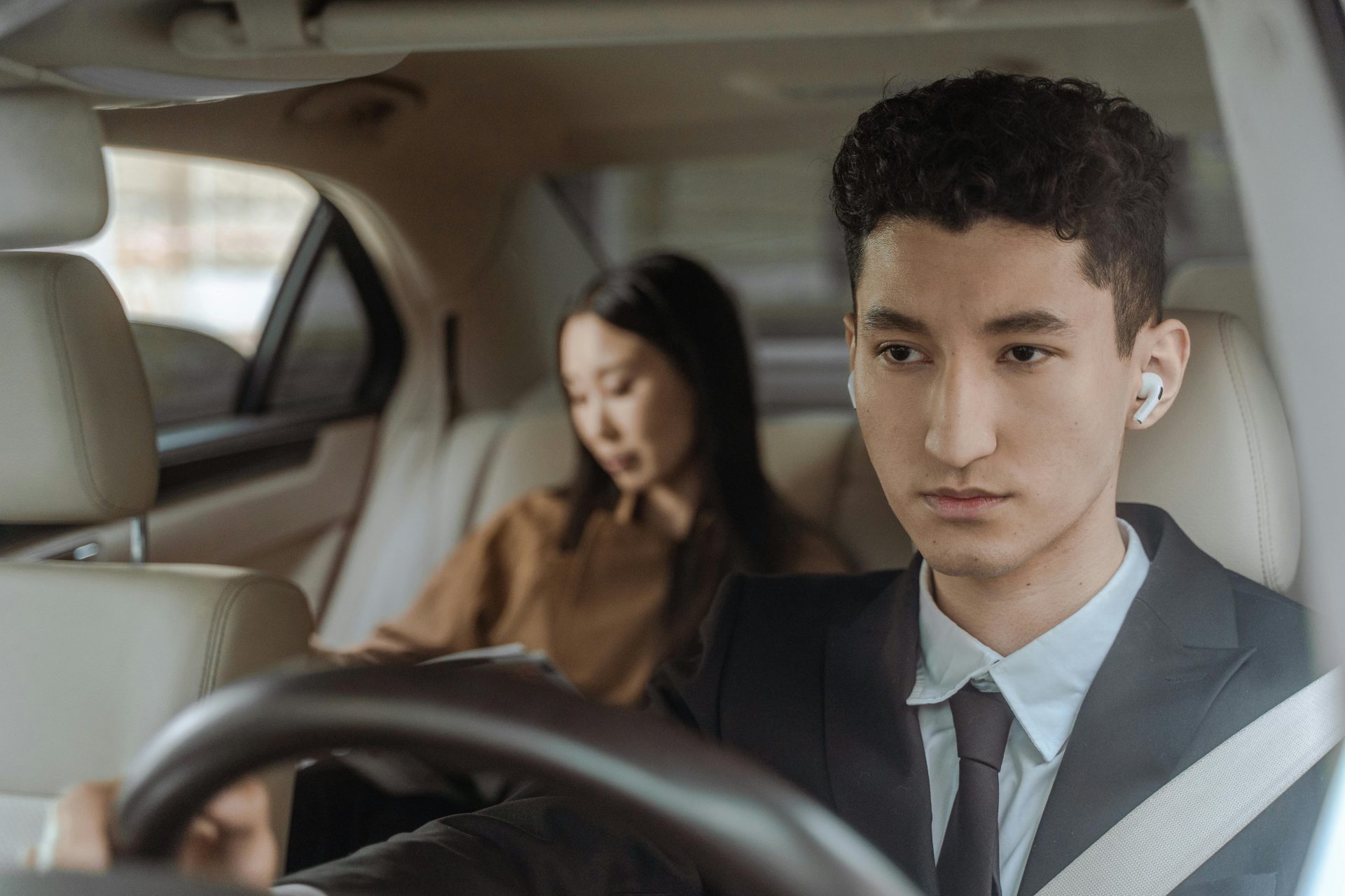 Junger, professioneller Fahrer im Anzug und mit Ohrhörer im Innenraum eines Luxuswagens mit weiblicher Beifahrerin.